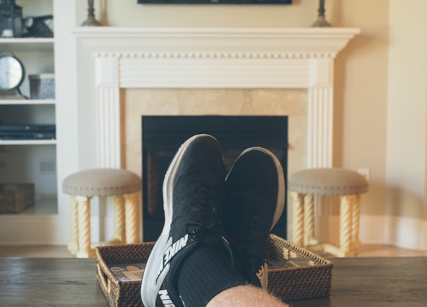 A person is relaxing in a living room with feet up on a wooden table. A TV mounted above a fireplace is displaying a popular streaming platform. Shelves on either side of the fireplace hold decorative items. A smartphone can be seen on the table.