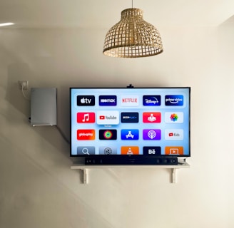 A modern living room features a wall-mounted flat-screen television displaying multiple streaming app icons, such as Apple TV, HBO Max, Netflix, Disney+, and Prime Video. Below the TV is a soundbar sitting on a small white shelf. A wicker pendant light hangs from the ceiling above, and the wall is painted a neutral color.